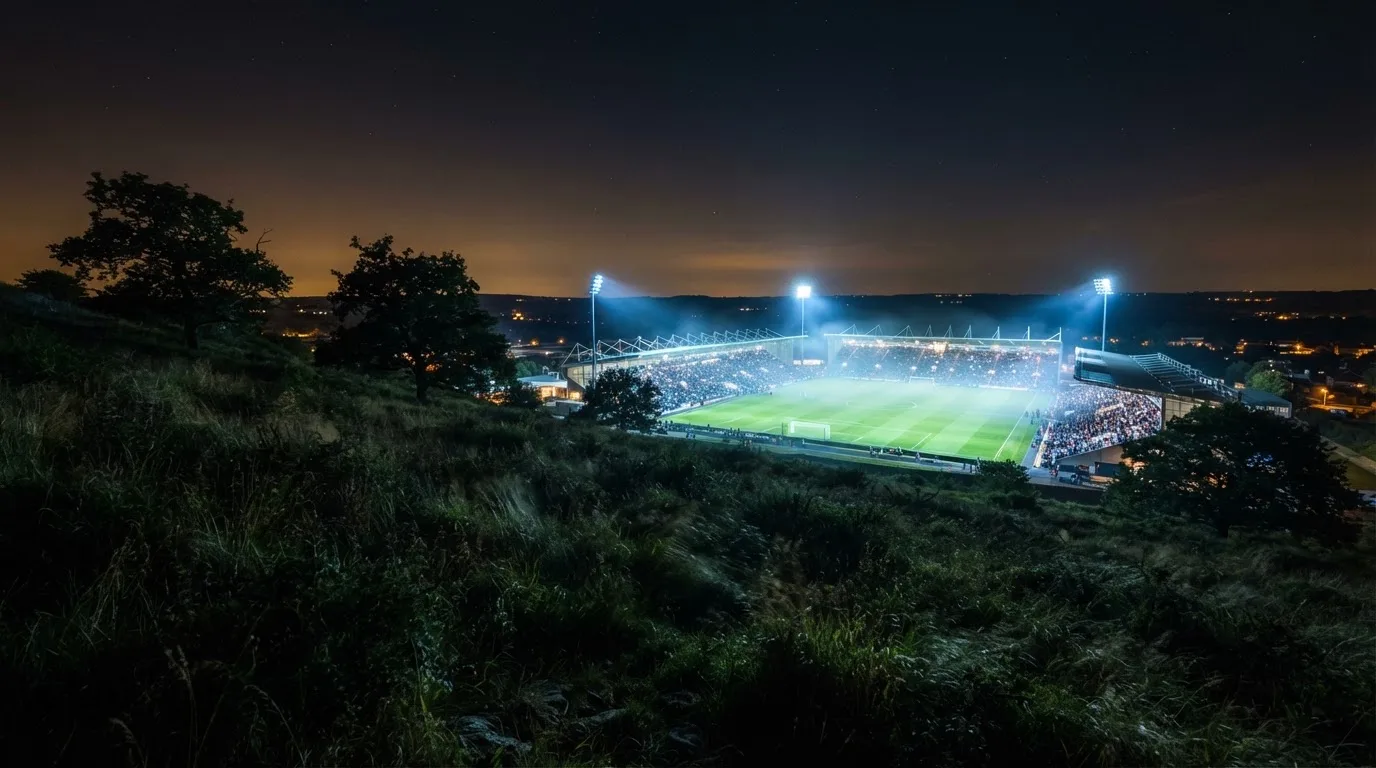 Ein Stadion unter Flutlicht bei Nacht, von einem Hügel aus der Ferne betrachtet, mit Dunst über dem Spielfeld.