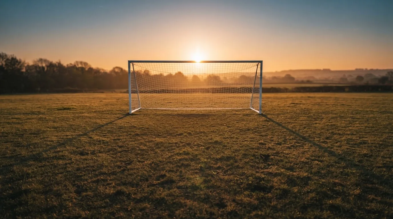 Ein Fußballtor mit Netz bei Sonnenuntergang auf einem leeren Platz, die Schatten der Torpfosten liegen lang auf dem Gras.