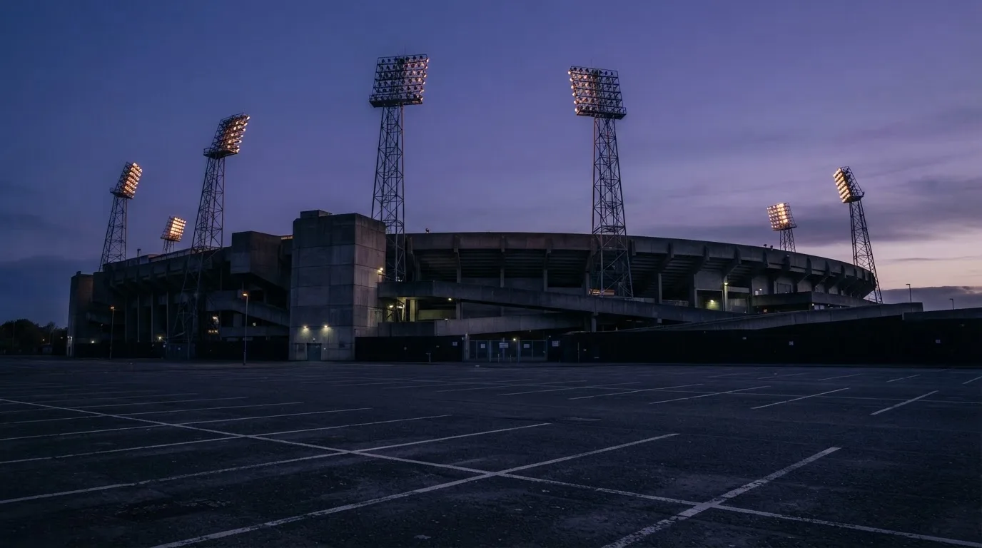 Ein Fußballstadion von außen in der Dämmerung, hohe Flutlichtmasten vor einem violetten Himmel, davor ein leerer Parkplatz.