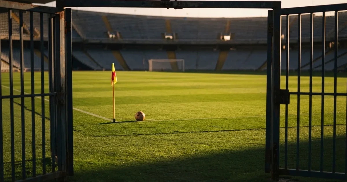Blick durch ein Stadiontor auf einen grünen Rasen im Abendlicht, ein Ball ruht an der Eckfahne, die Tribünen sind leer.