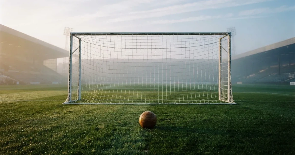 Ein Fußballtor mit gespanntem Netz steht auf einem leeren Rasenplatz, davor ein einzelner Ball, Morgendunst liegt über dem Feld.