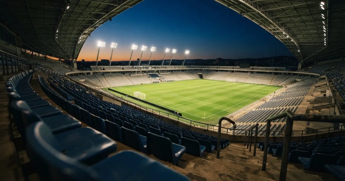 Leere Sitzreihen eines großen Stadions unter Flutlicht, dahinter die ferne Sicht auf den hell beleuchteten Rasen nach Sonnenuntergang.