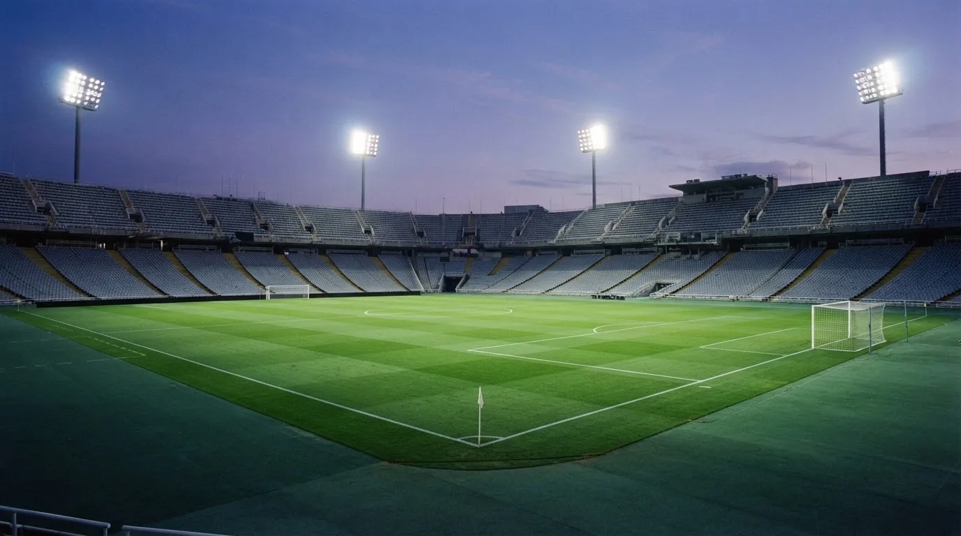 Eine leere Stadiontribüne unter Flutlicht aus der Ferne betrachtet, die Rasenfläche leuchtet grün in der Dämmerung.