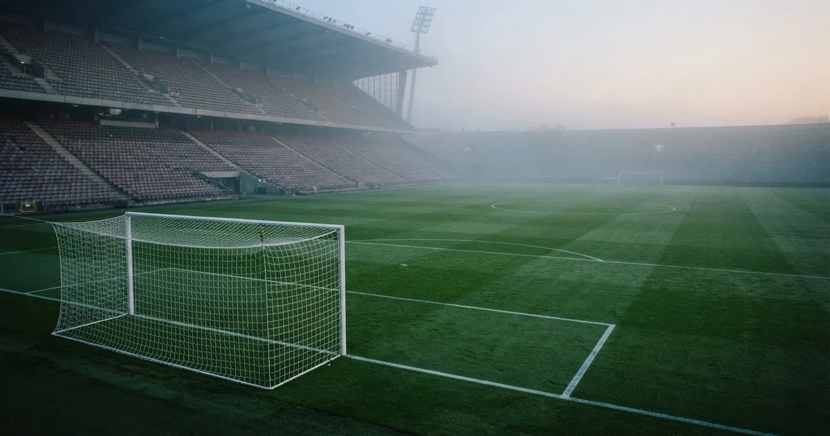 Ein Fußballplatz mit gepflegtem Rasen und einem Tor, über dem Spielfeld liegt Morgennebel, im Hintergrund eine leere Tribüne.