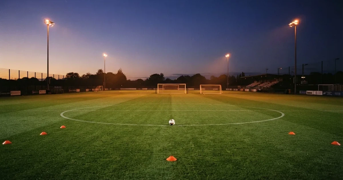 Eine Flutlichtanlage ragt über einen Trainingsplatz, auf dem Rasen liegen verstreut Hütchen und ein Ball, am Himmel die Abenddämmerung.