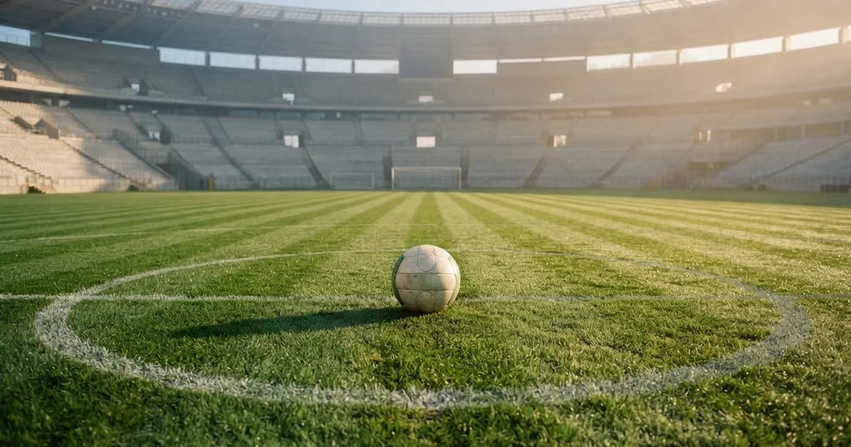 Ein Fußball liegt auf dem Mittelkreis eines Stadionrasens am frühen Morgen, im Hintergrund leere Ränge ohne erkennbare Beschriftung.