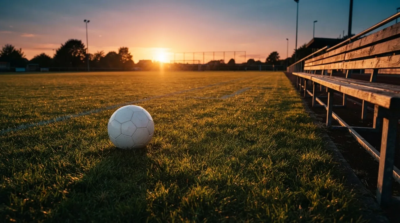 Ein Fußball auf einem Rasenplatz im Gegenlicht der tiefstehenden Sonne, am Rand eine leere Ersatzbank.
