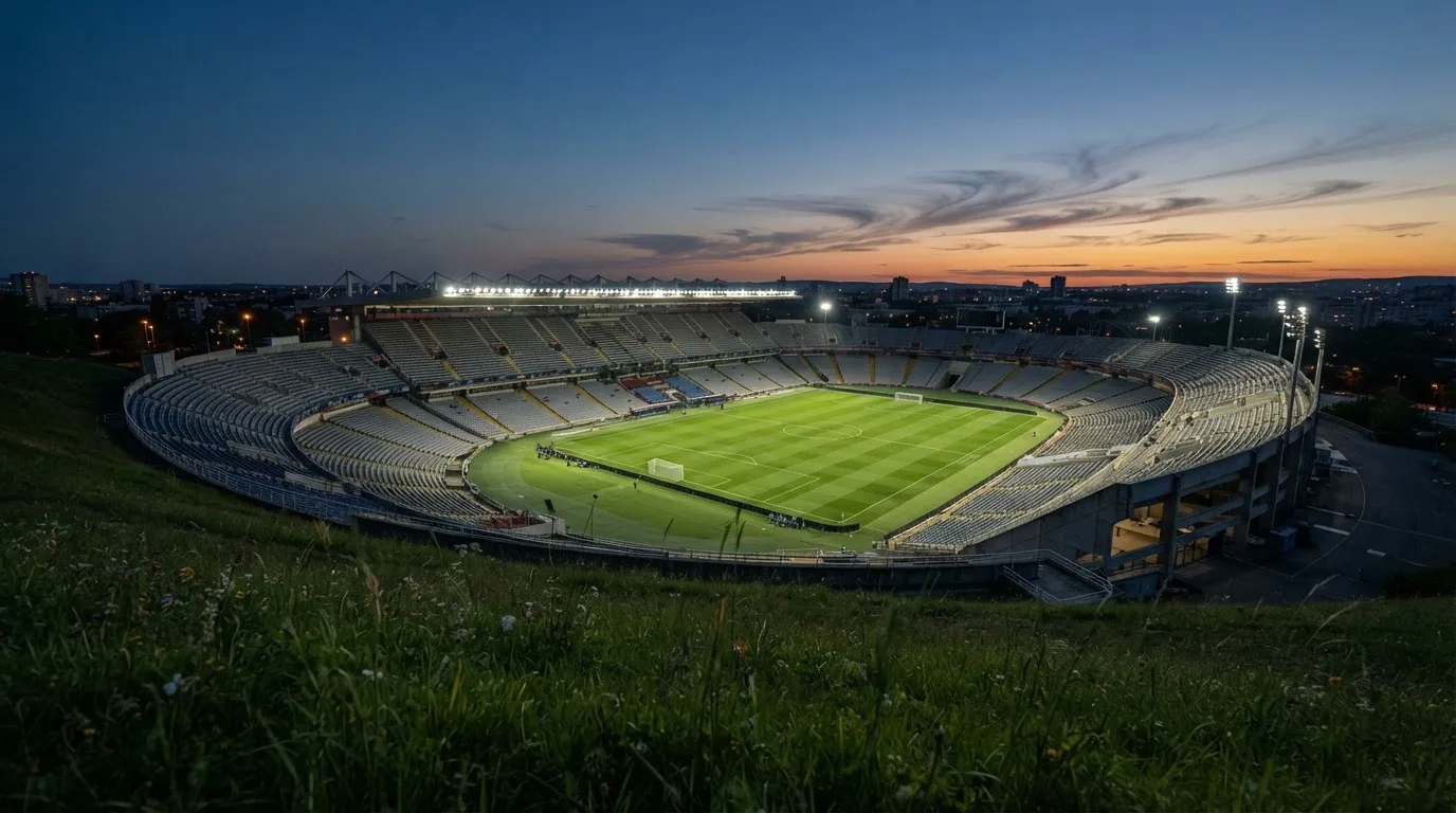 Ein leeres Stadion mit hell erleuchteten Rängen in der Abenddämmerung, von einem Hügel aus fotografiert.