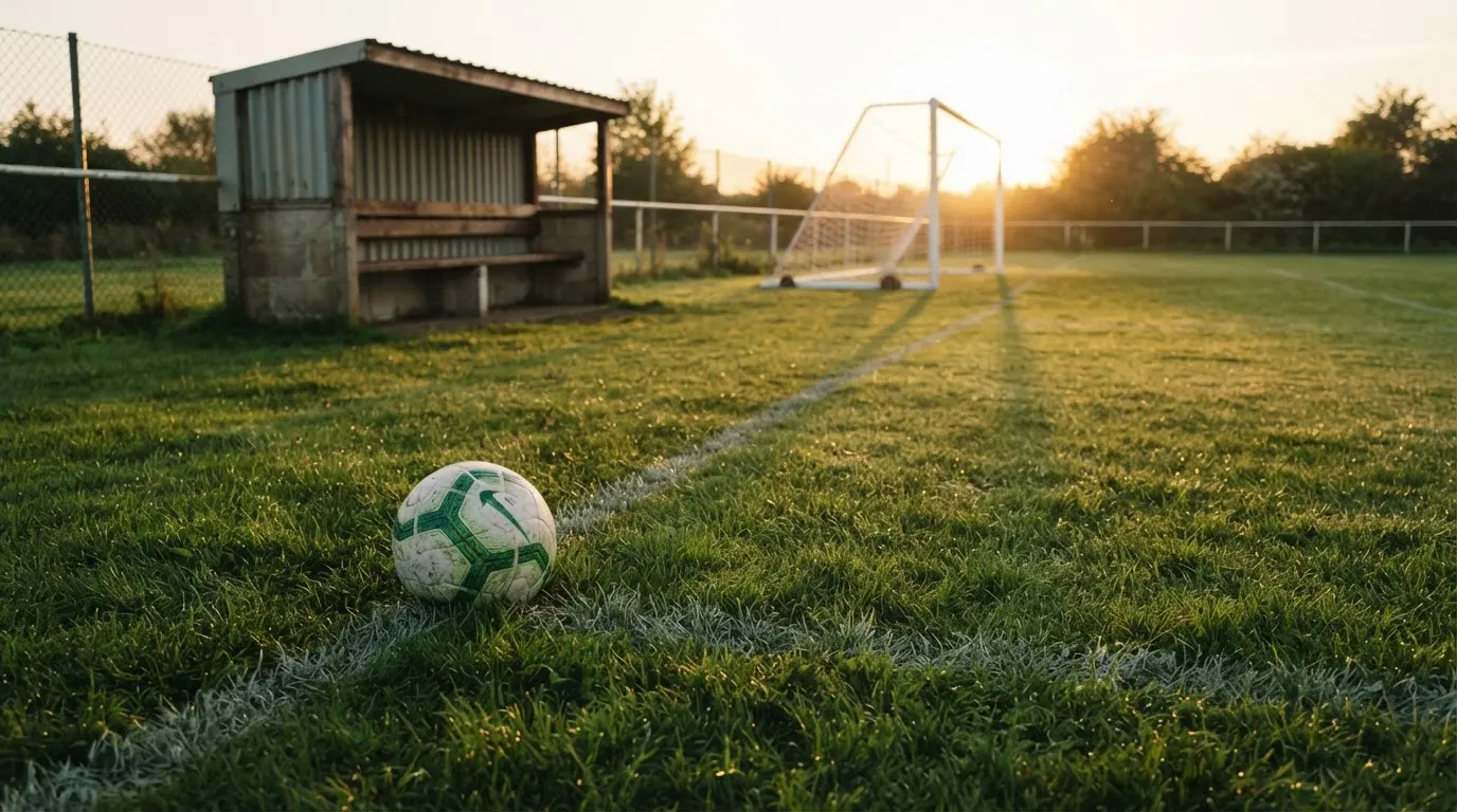 Ein Fußball liegt im Gras am Spielfeldrand, dahinter eine leere Bank und ein Tor im Gegenlicht.