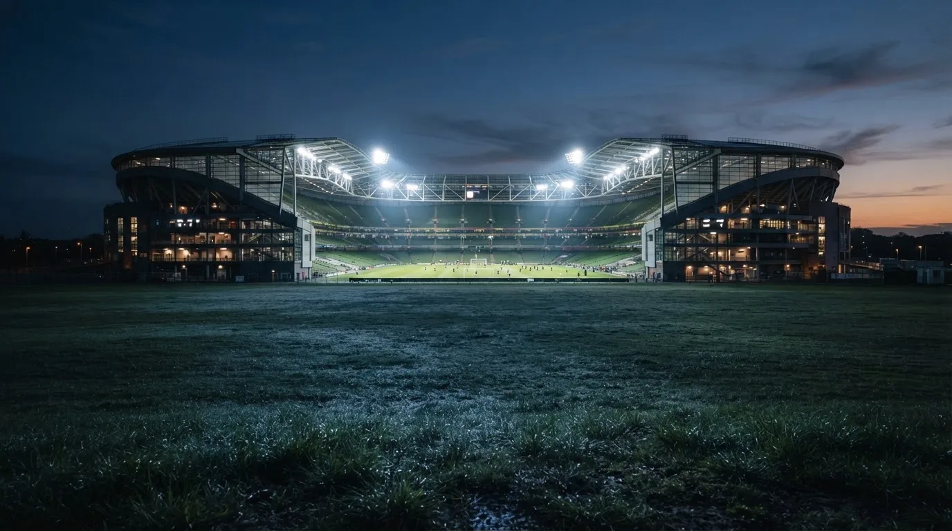 Ein Stadion unter Flutlicht aus der Ferne, davor ein stilles Feld im Dämmerlicht.