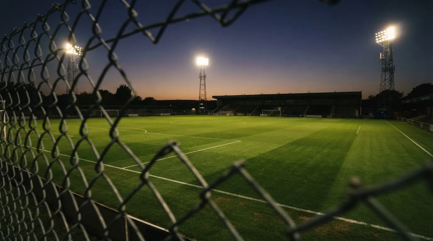 Eine Flutlichtanlage über einem leeren Sportplatz am Abend, im Vordergrund ein Maschendrahtzaun.