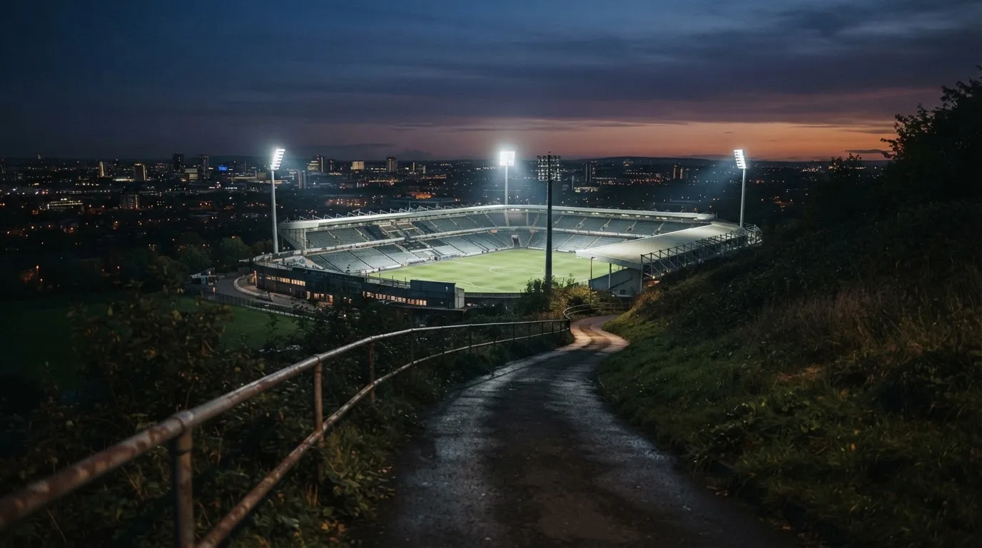 Ein Stadion unter Flutlicht, von einer Anhöhe aus gesehen, davor ein stiller Weg in der Dämmerung.