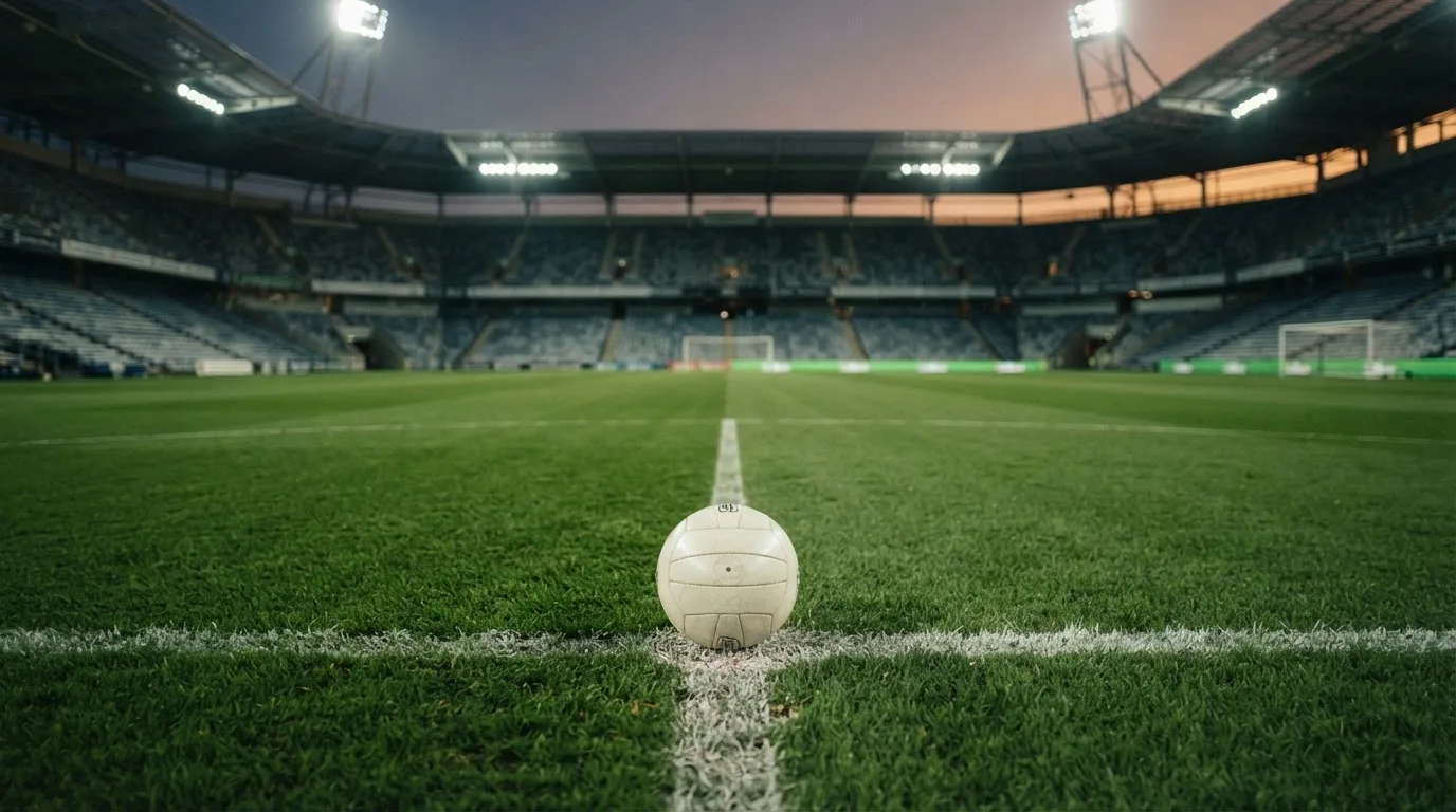 Ein Fußball liegt auf dem Anstoßpunkt eines leeren Rasenplatzes, im Hintergrund ein Stadion unter Flutlicht in der Abenddämmerung.