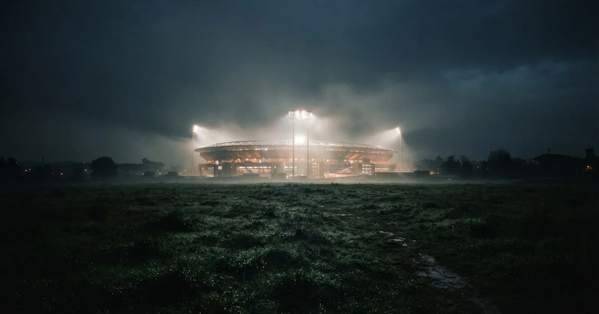 Ein Stadion unter Flutlicht, aus großer Entfernung über eine dunkle Wiese fotografiert, am Himmel zieht Nebel auf.