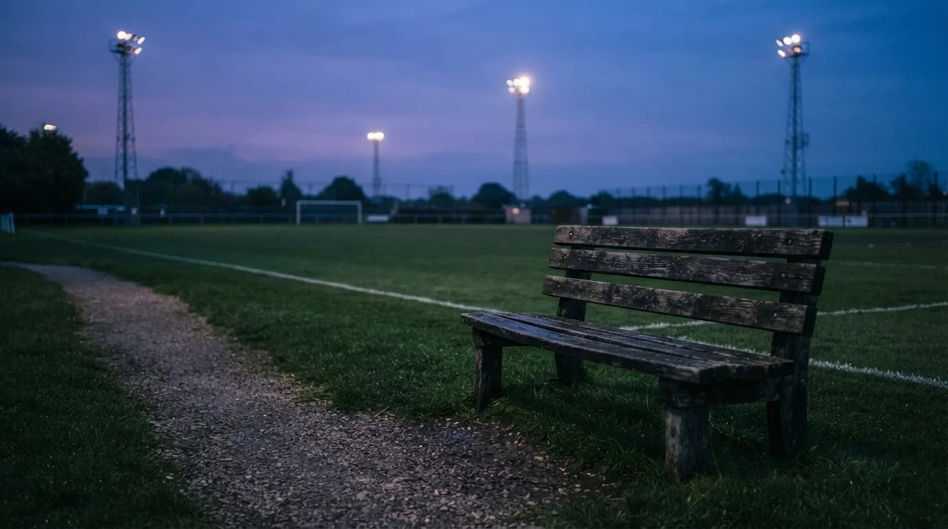 Eine leere Parkbank am Rand eines Fußballfelds in der Abenddämmerung, im Hintergrund ragen dunkle Flutlichtmasten in den Himmel.