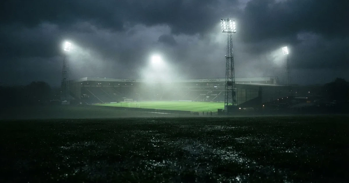 Ein Stadion unter Flutlicht, aus großer Entfernung über eine dunkle Wiese fotografiert, am Himmel zieht Nebel auf.