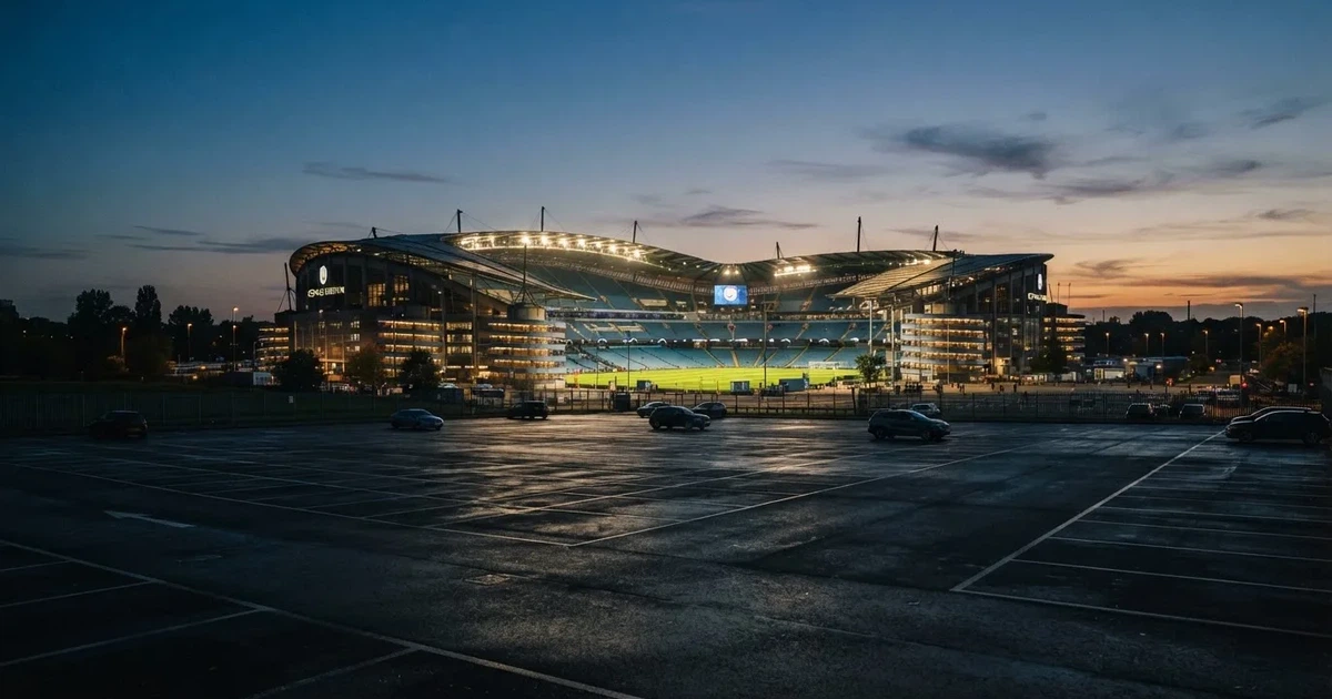 Ein Fußballstadion unter Flutlicht aus großer Entfernung, davor ein dunkler Parkplatz und ein heller Abendhimmel.
