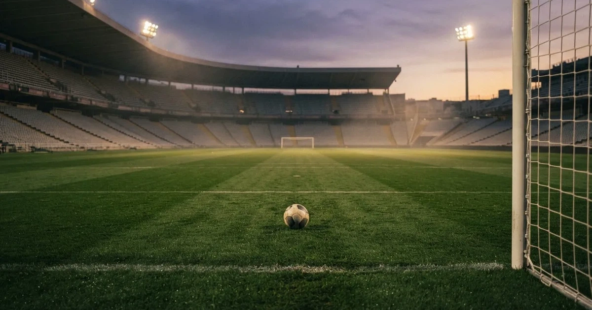 Ein Fußballplatz mit frisch gemähtem Rasen im Abendlicht, ein Ball liegt im Strafraum, im Hintergrund unscharf eine leere Tribüne.