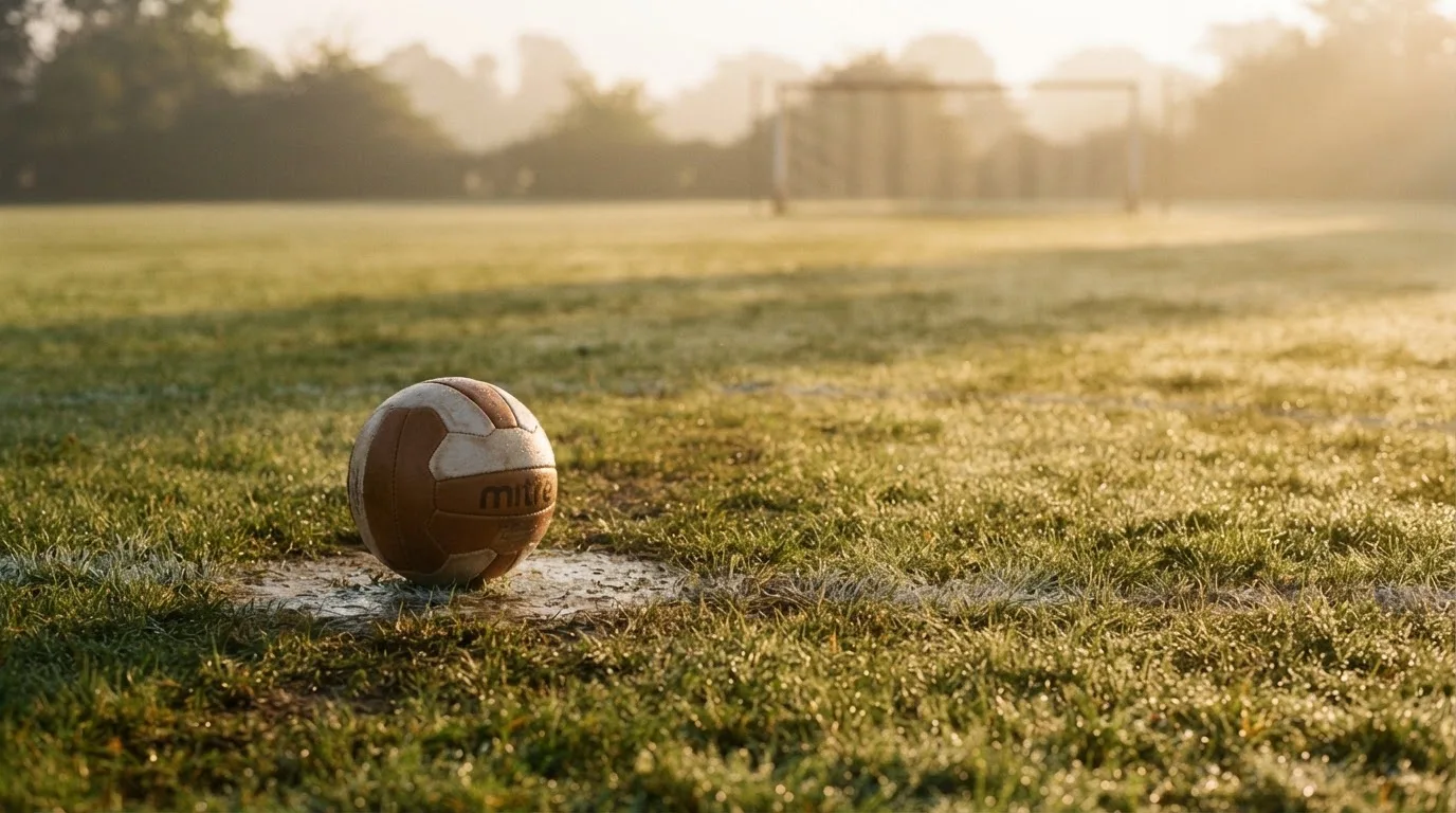 Ein Fußball ruht auf dem Mittelkreis eines leeren Rasenplatzes in der Morgendämmerung, Tau liegt auf dem Gras.