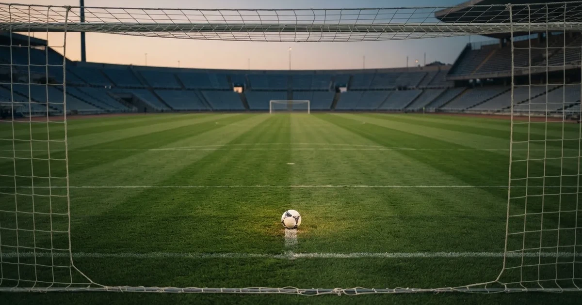 Ein Fußballplatz mit frisch gemähtem Rasen im Abendlicht, ein Ball liegt im Strafraum, im Hintergrund unscharf eine leere Tribüne.