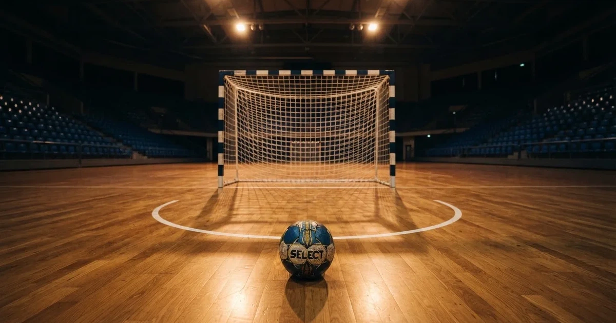 Ein Handball liegt auf dem Hallenboden vor einem Tor mit Netz, die leere Halle wird von Flutlicht beleuchtet.