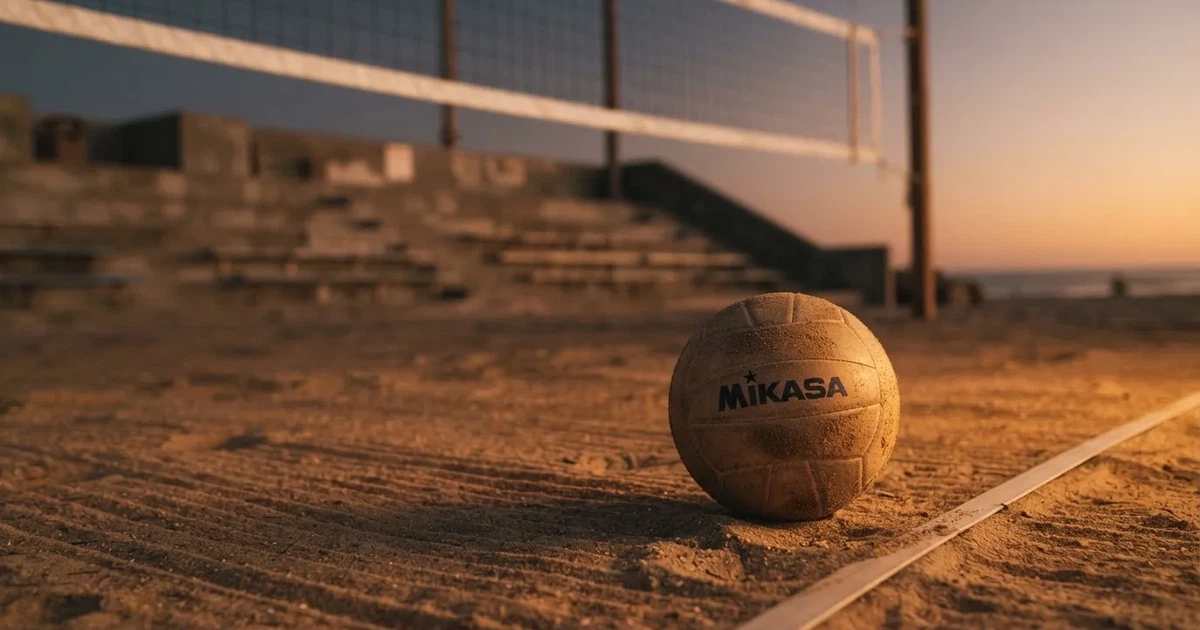 Ein Volleyball liegt auf dem Sand eines Beachvolleyball-Feldes neben dem gespannten Netz, im Hintergrund eine leere Tribüne bei Abendlicht.