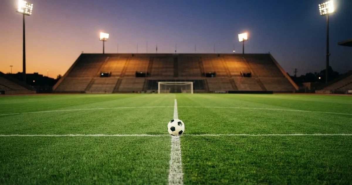 Ein Fußball liegt auf dem Anstoßpunkt eines leeren Rasenplatzes, im Hintergrund eine Tribüne und Flutlichtmasten in der Abenddämmerung.