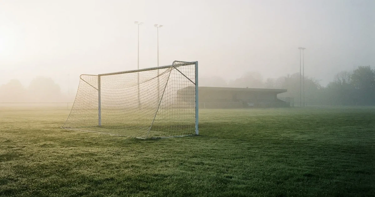 Ein Fußballtor mit Netz steht im Morgennebel auf einem leeren Rasenplatz, im Hintergrund die Silhouette einer kleinen Tribüne.