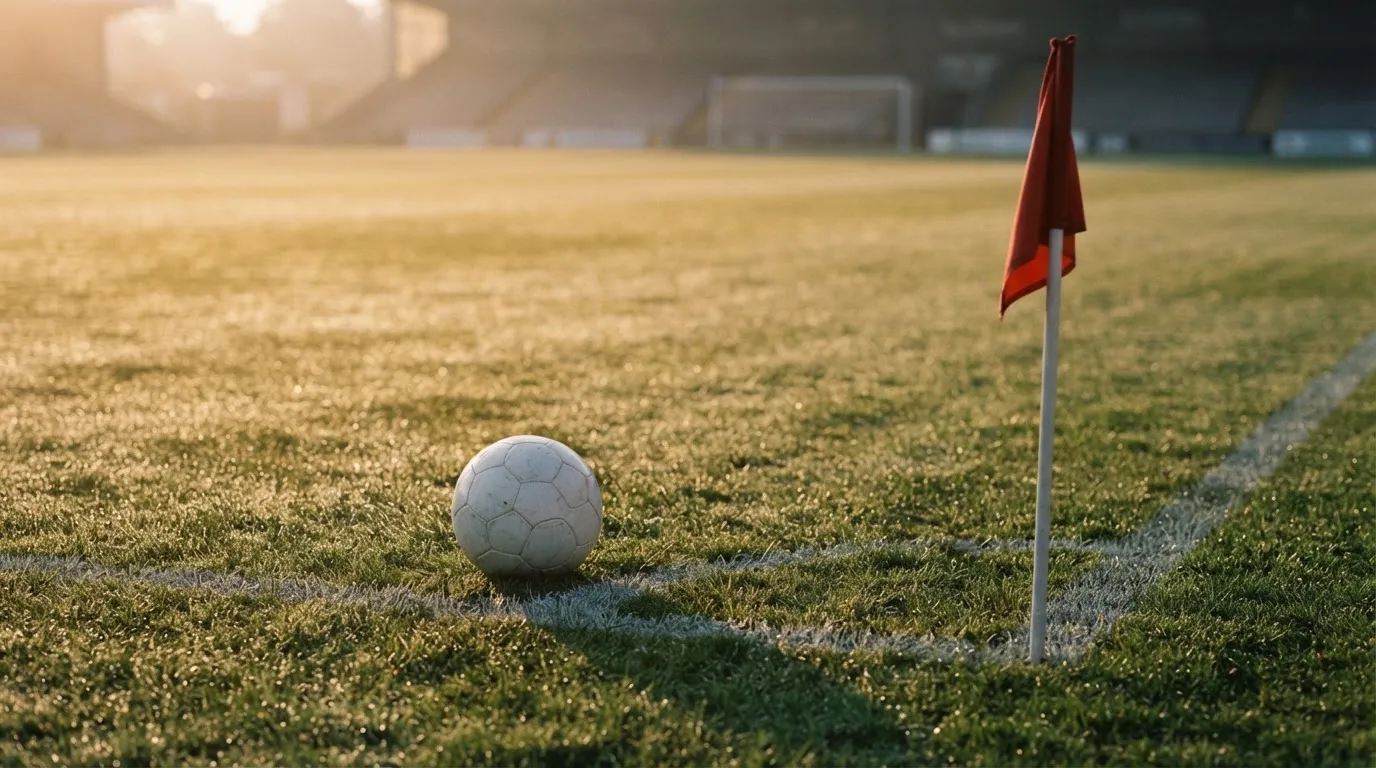 Ein Fußball liegt auf Rasen neben einer Eckfahne ohne Aufdruck auf einem leeren Platz im Morgenlicht.