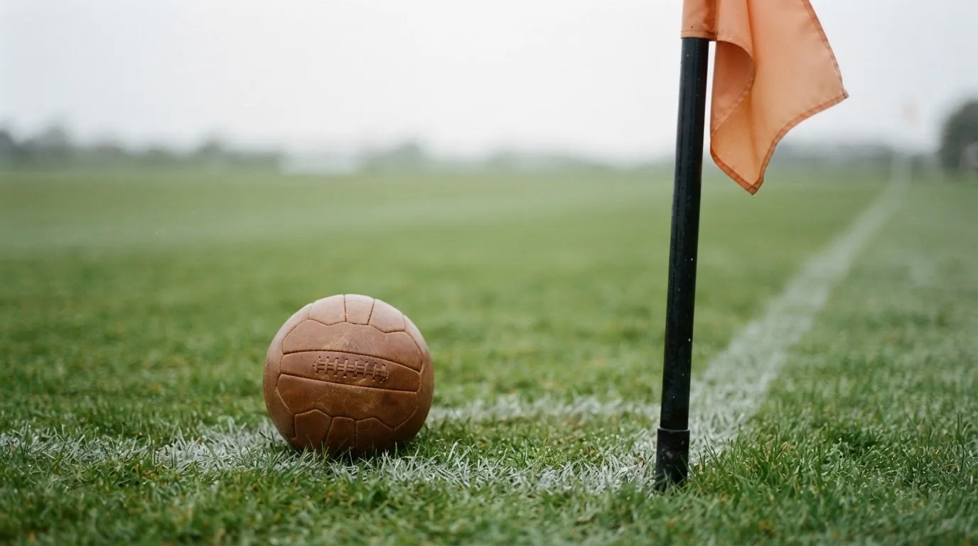 Ein Fußball liegt neben einer Eckfahne auf kurz gemähtem Rasen unter bewölktem Himmel.