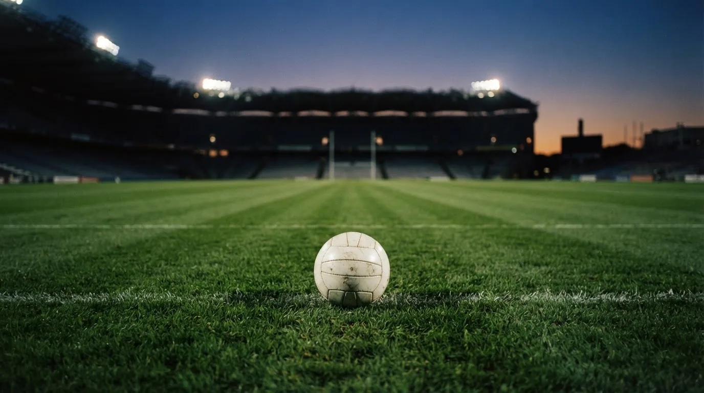 Ein Fußball liegt auf einem Rasenplatz in der Dämmerung, im Hintergrund ein ferner Stadionrand unter Flutlicht.
