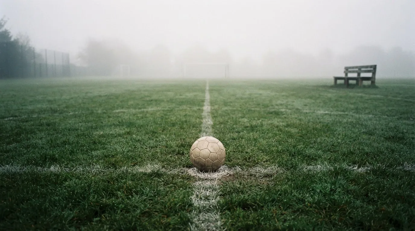 Ein Fußball liegt in der Mitte eines leeren Rasenplatzes im Morgennebel, am Rand steht eine einzelne Auswechselbank.